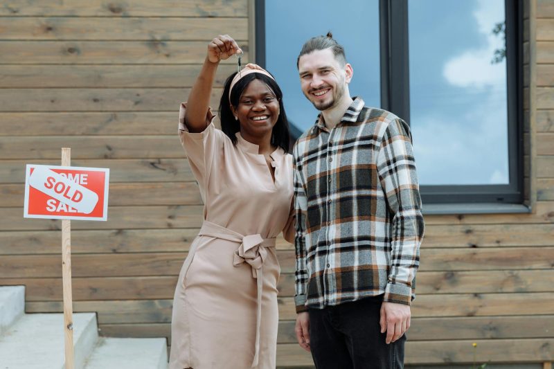 Young couple joyfully holds house keys in front of their new home with a sold sign.