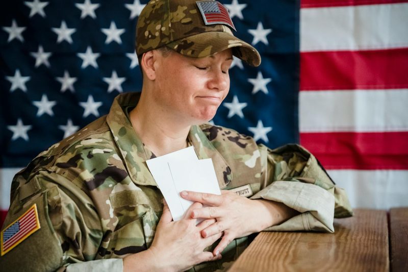 A soldier in uniform holds envelopes in front of a large American flag.
