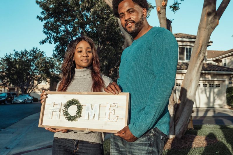 Smiling couple holding a "Home" sign in their neighborhood, celebrating new beginnings.