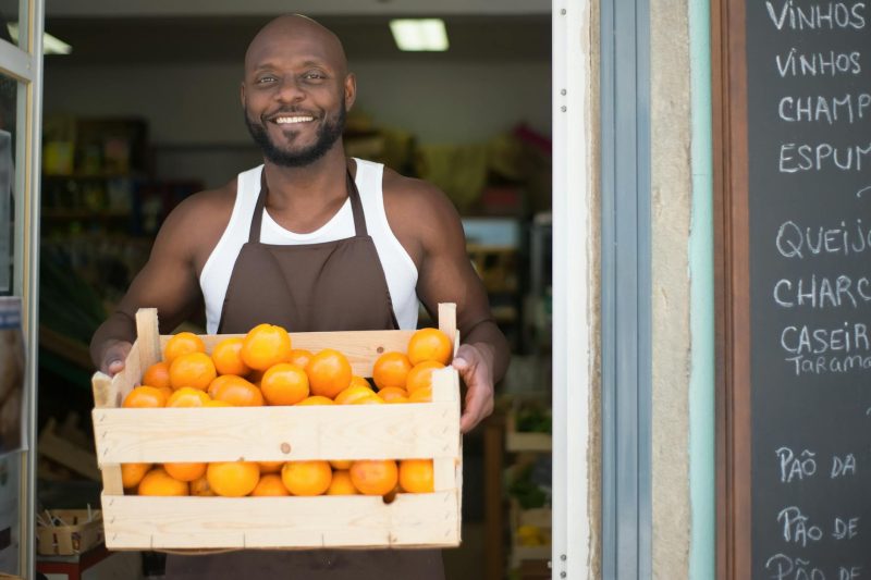 Vendor smiling while holding a crate of oranges in a colorful local store in Portugal.