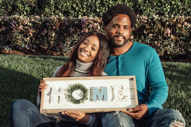Smiling couple sitting on a lawn, happily holding a 'Home' sign to celebrate their new home.