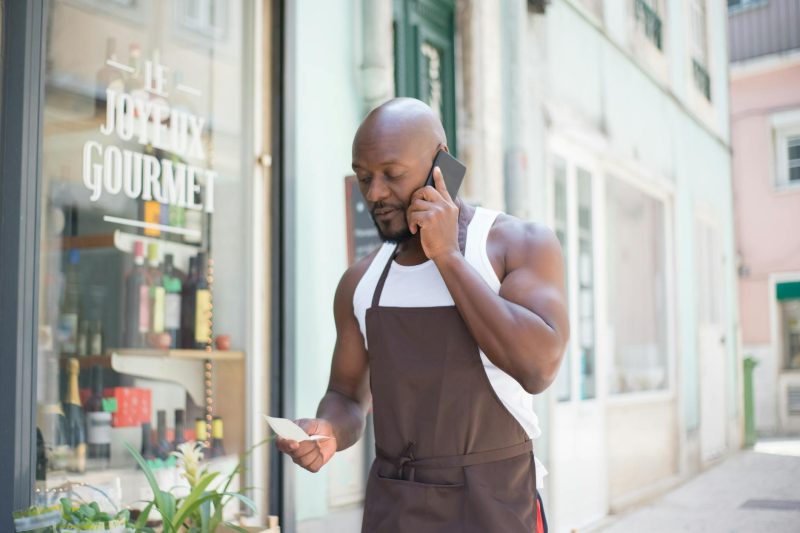 Man in apron making a business call outside a gourmet store in Portugal, reviewing notes.