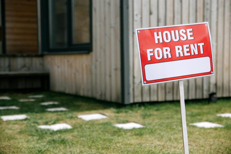 Modern wooden house in a green yard with a prominent 'House for Rent' sign.