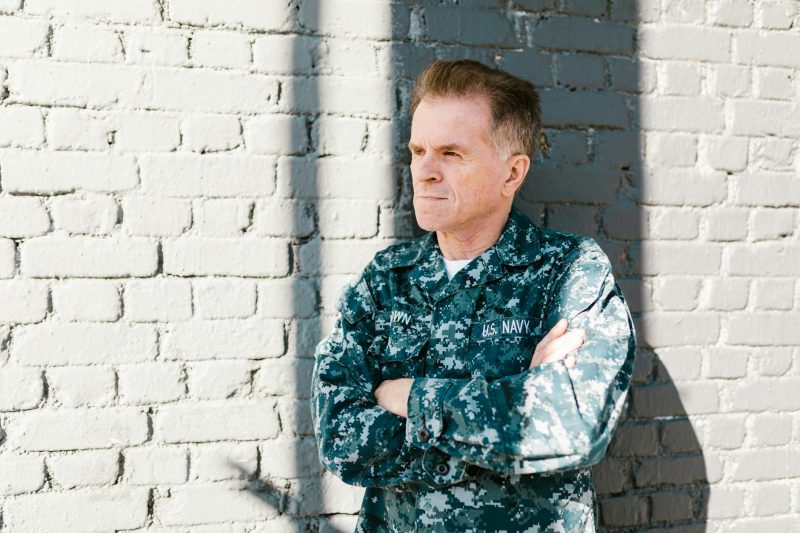 Senior US Navy veteran in uniform leans against a sunlit brick wall, conveying a sense of pride and reflection.