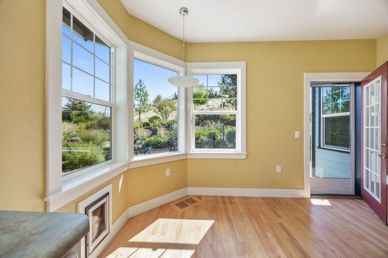 Home 3 Spacious room with wooden floors, large windows showcasing garden views, illuminated by sunlight.