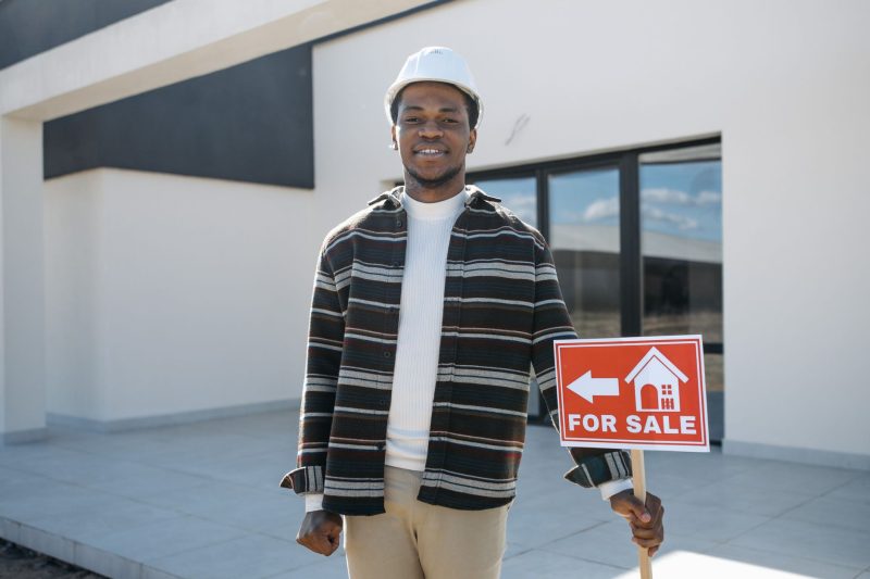 Home 3 Young black male realtor smiling while holding a for sale sign in front of a modern house.